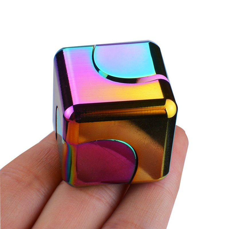 Square Fidget Cube shown as a metallic rainbow-colored dice cube with iridescent finish, featuring smooth curved edges and geometric patterns. The compact stress relief toy displays vibrant holographic effects in pink, blue, yellow, and purple tones, held between fingers to demonstrate its size.