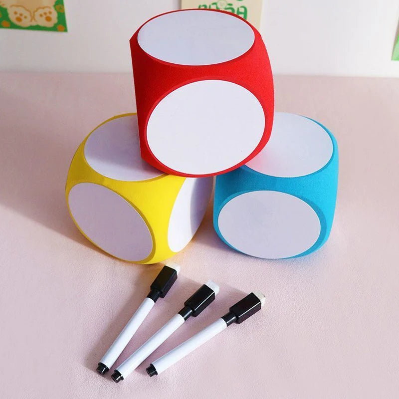 Blank Dice Write-On Cube displayed in red, yellow, and blue colors with white writable surfaces, shown with three black dry-erase markers for customization. The dice are stacked in a pyramid formation on a pink surface.