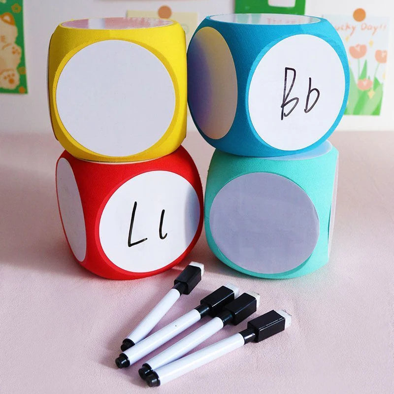 Blank Dice Write-On Cube displayed in four vibrant colors - yellow, blue, red, and mint green - with white circular faces for customization, shown alongside black dry-erase markers. One die shows handwritten letters demonstrating the writable surface.