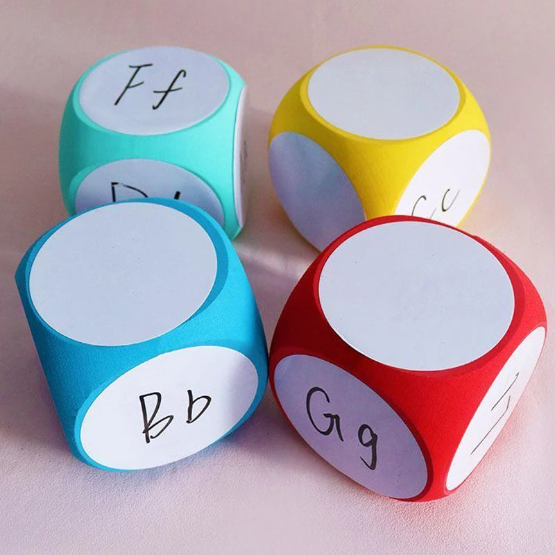 Blank Dice Write-On Cube displayed in four vibrant colors - red, yellow, turquoise, and mint green - with white writable surfaces and handwritten letters visible on some faces, arranged on a pink background, showcasing their customizable educational and gaming potential.