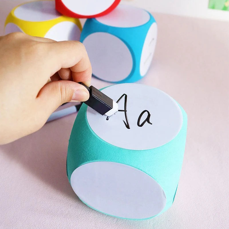 Blank Dice Write-On Cube shown being customized with a marker, featuring a teal colored foam die with white writable surfaces on each face. Multiple colorful dice visible in background demonstrating the product's variety and customization potential.