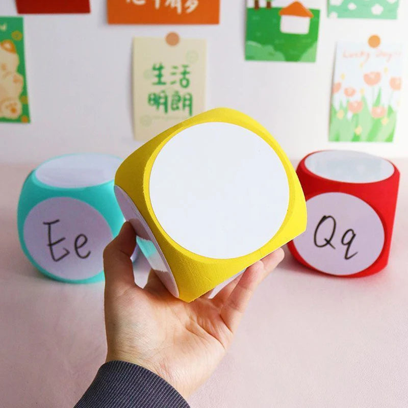 Blank Dice Write-On Cube shown in yellow, with red and turquoise cubes in background. Hand holding yellow cube demonstrates size and writable white surface. Educational cards visible on wall behind cubes. Letters Ee and Qq visible on companion dice.