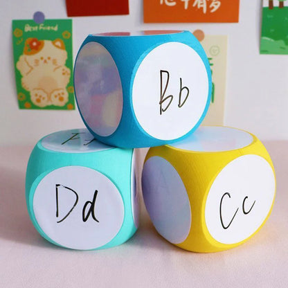 Blank Dice Write-On Cube set featuring three colorful dice in blue, mint, and yellow with handwritten letters bb, Dd, and Cc on white circular faces, displayed against a light background with decorative cards on the wall.