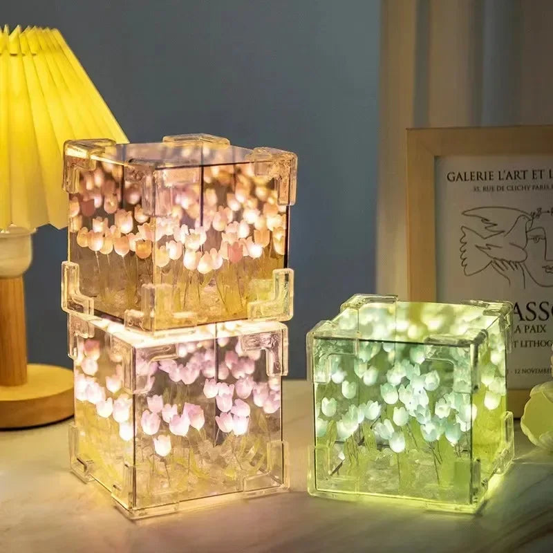 Creative Tulip Flower Sea Cube night light featuring transparent cube-shaped lamps with illuminated tulip flowers creating a dreamy underwater effect. Two stacked cubes emit a warm golden glow with pink flowers, while a separate cube glows in soft green, displayed on a table alongside a yellow lamp and artwork.