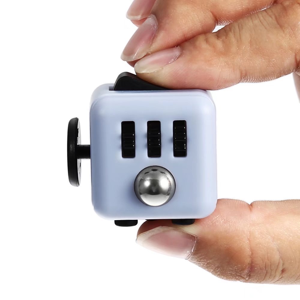 Fidget Cube Toy shown in light blue color with black buttons and silver ball, being held between fingers to demonstrate its compact size. Features multiple clickable surfaces for stress relief and sensory stimulation.