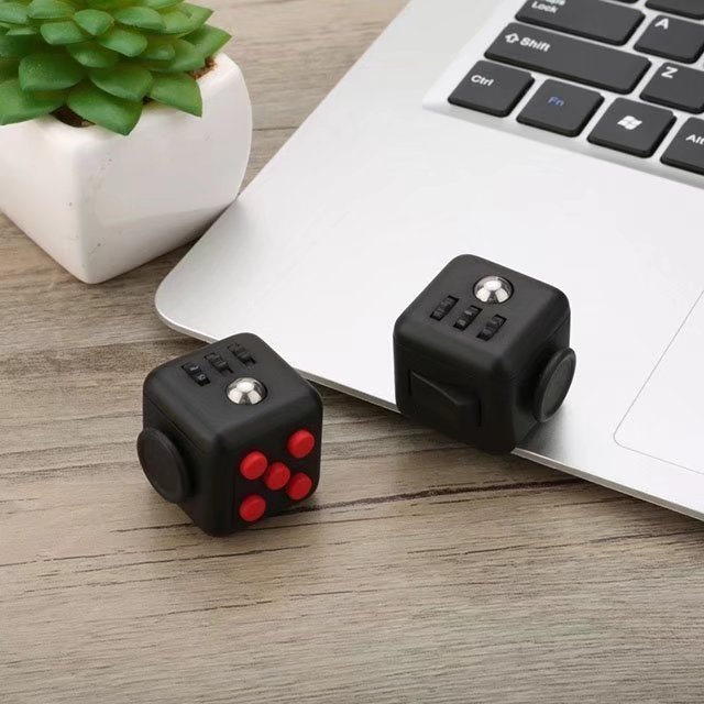 Fidget Cube Toy displayed next to laptop and small plant, featuring black cubes with red buttons and interactive surfaces for stress relief and entertainment. Compact 3.3cm size shown in desk setting with modern minimalist aesthetic.