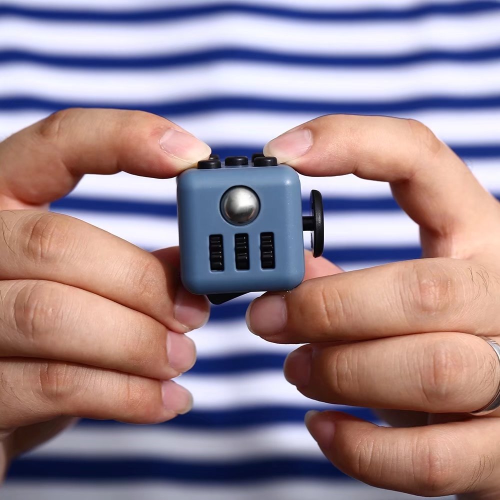 Fidget Cube Toy shown in blue color being held between fingers against striped background, featuring multiple tactile buttons and switches for stress relief and sensory stimulation. Small portable anxiety relief device demonstrating size and functionality.