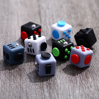 Fidget Cube Toy collection displayed on wooden surface, featuring multiple cubes in various colors including black, white, and blue, each with different interactive buttons, switches, and tactile elements for stress relief and entertainment